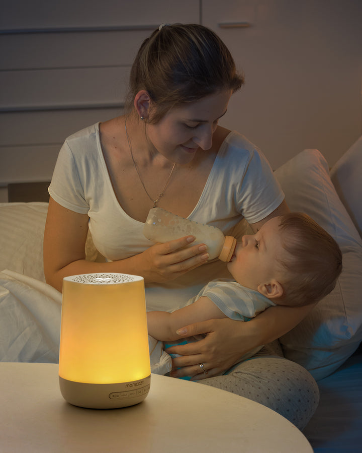 Mother feeding baby with a bottle next to glowing smart baby sound machine, illustrating a peaceful nighttime routine.