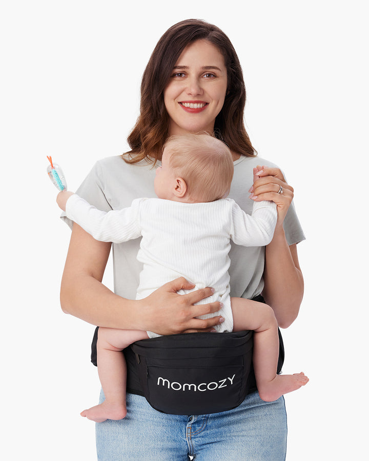 Mother holding a baby on black Momcozy baby hip seat carrier, baby holding a toy, showcasing the product's functionality
