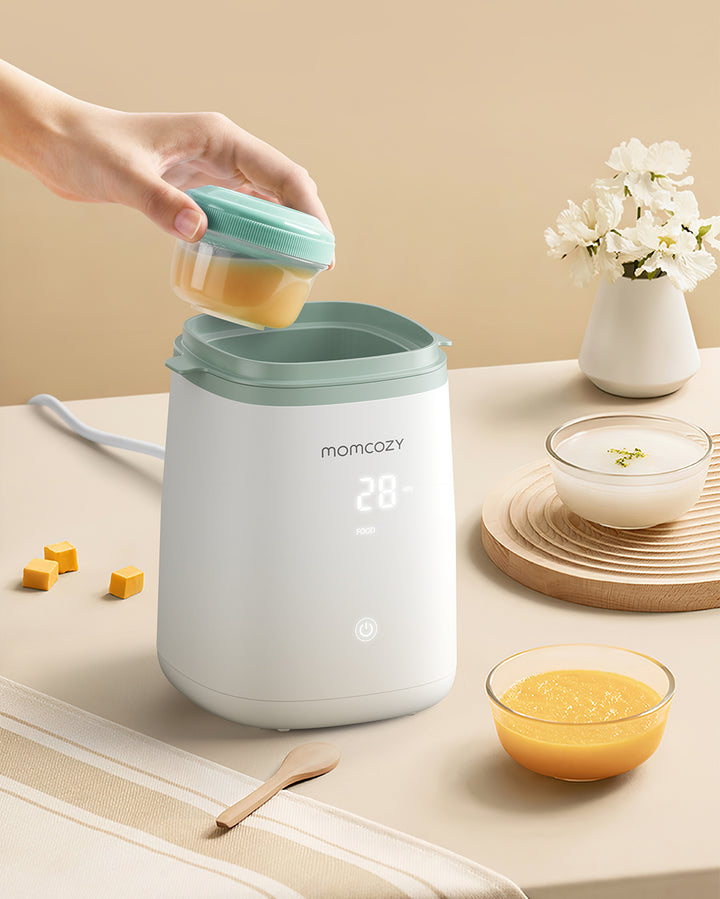 Momcozy 6-in-1 Fast Baby Bottle Warmer with digital display showing '28', a person placing a container with baby food inside the warmer. Surrounding items include a small bowl of baby food, baby food cubes, and a vase with white flowers.