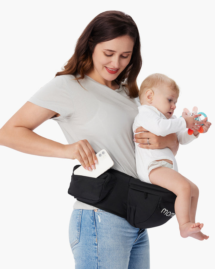 Woman holding baby in black Momcozy baby hip seat carrier with multiple pockets, placing a smartphone into one of the pockets, highlighting the carrier's functionality and convenience.