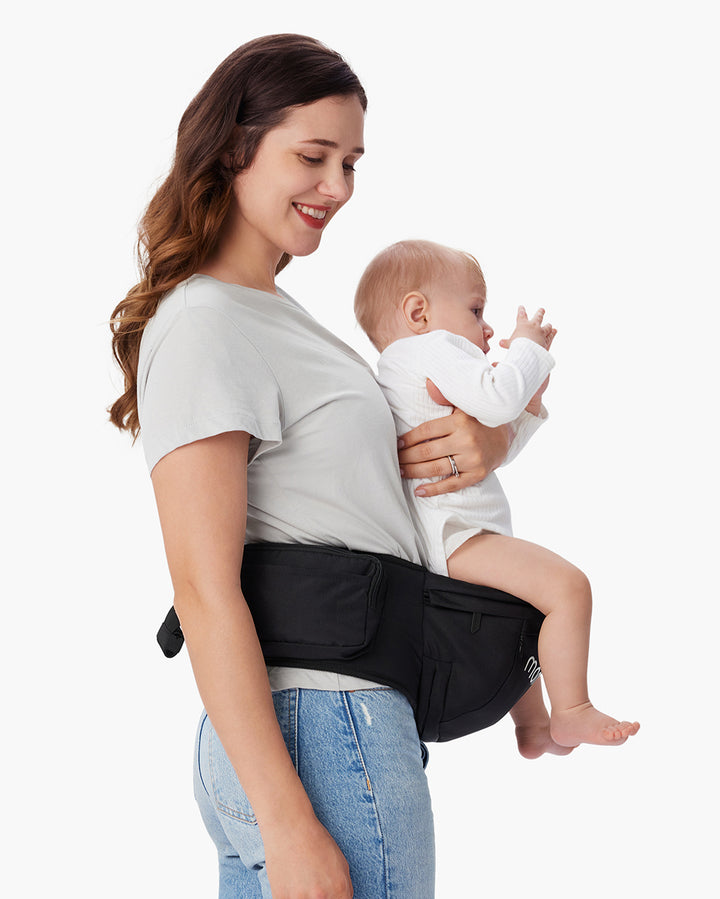 Mother wearing grey t-shirt and blue jeans carrying baby in white onesie using black baby hip seat carrier, showcasing comfort and convenience of the Momcozy baby hip seat carrier.