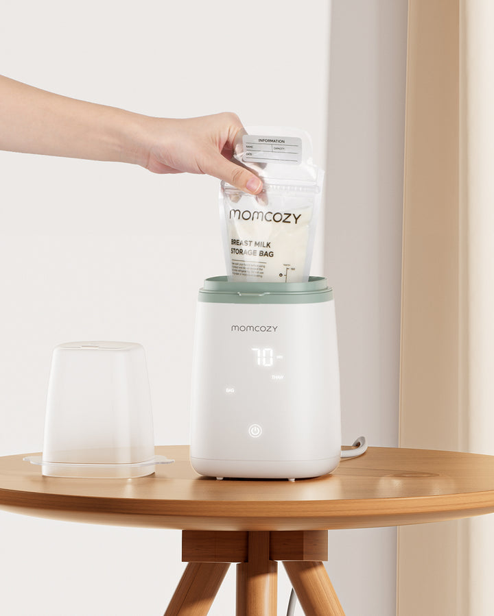 Hand placing a Momcozy breast milk storage bag into a Momcozy 6-in-1 Fast Baby Bottle Warmer on a wooden table. The warmer features a digital temperature display.