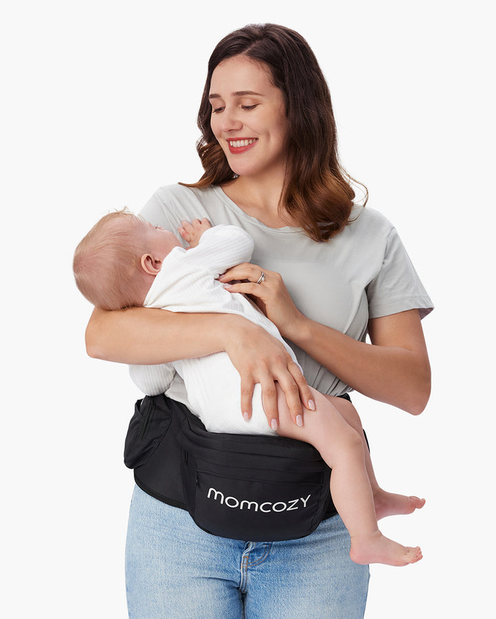 Mother holding baby using Momcozy baby hip seat carrier, smiling at her baby. The black carrier has 'Momcozy' printed in white on the front.
