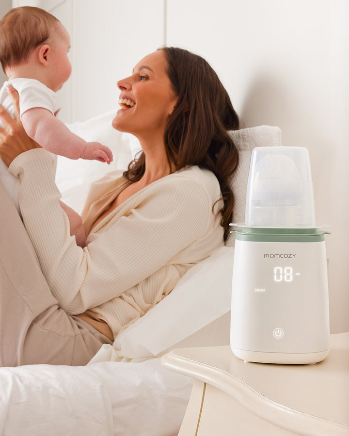 Mother holding baby on bed next to Momcozy 6-in-1 Fast Baby Bottle Warmer displaying '08' on LED screen, highlighting versatile functionality and convenience for mothers.