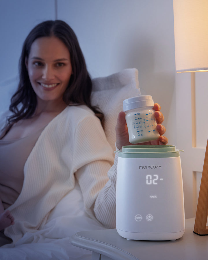 A woman using the Momcozy 6-in-1 fast baby bottle warmer, seen holding a baby bottle above the warmer which displays '02'.