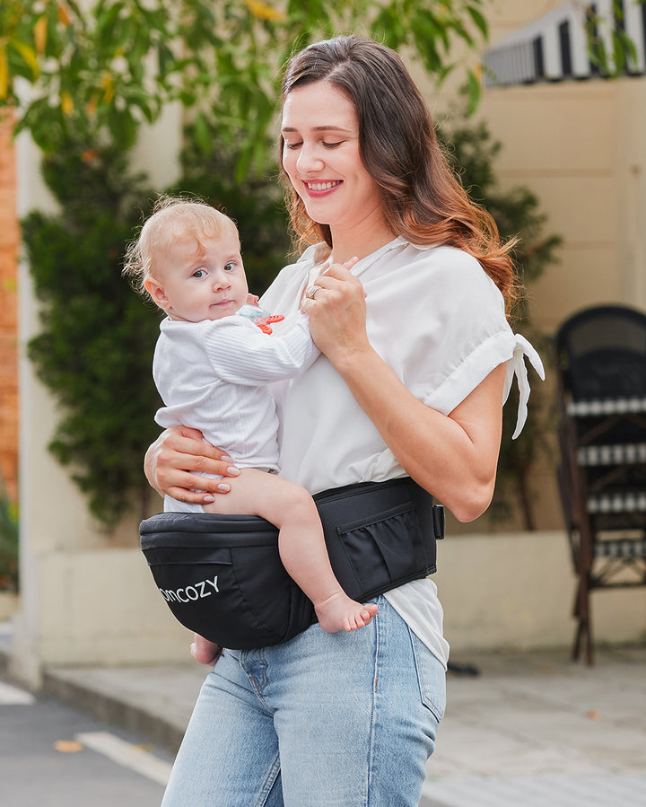 Mother holding baby in Momcozy 3D Belly Protector & EVA Massage Board-Baby Hip Seat Carrier outdoors.